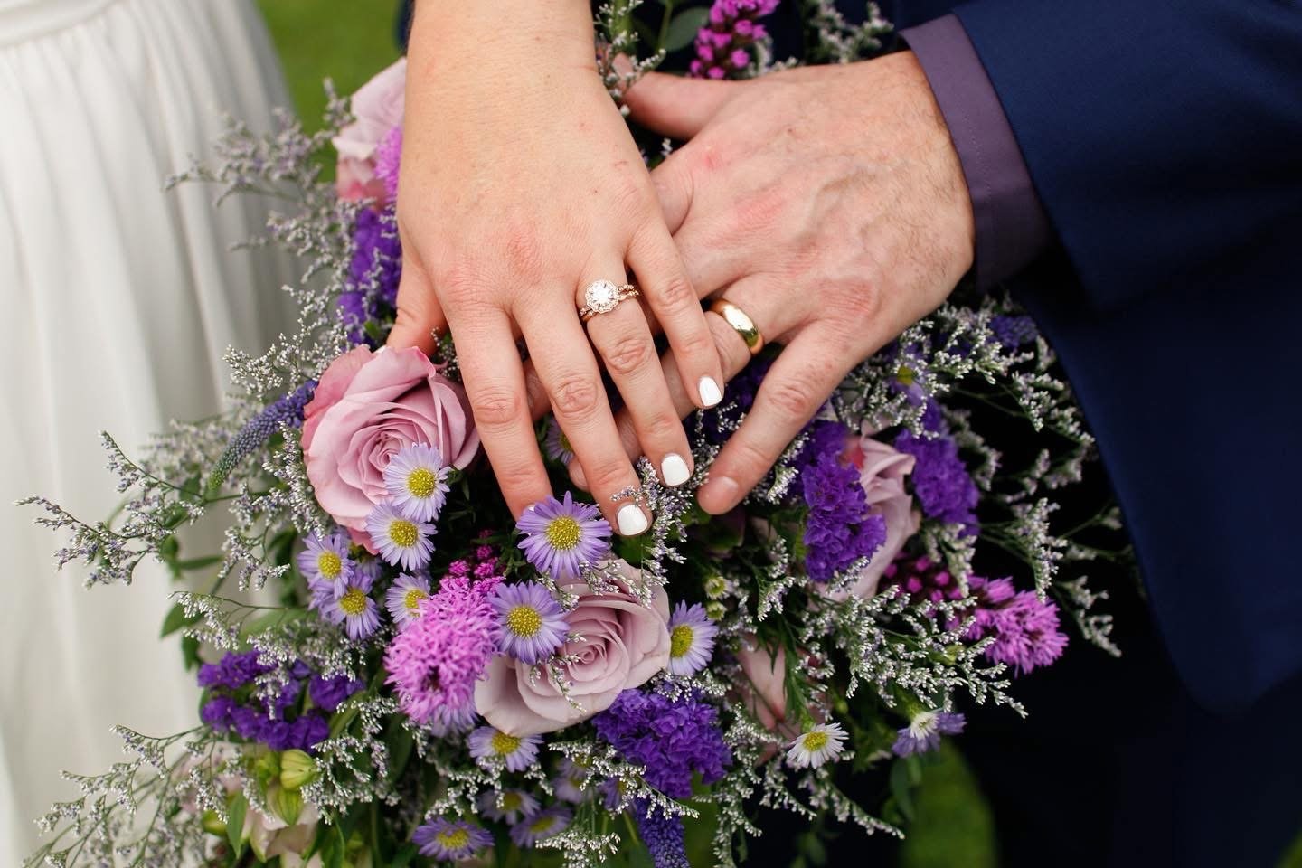 Newlyweds displaying their wedding rings over a purple and pink bridal bouquet